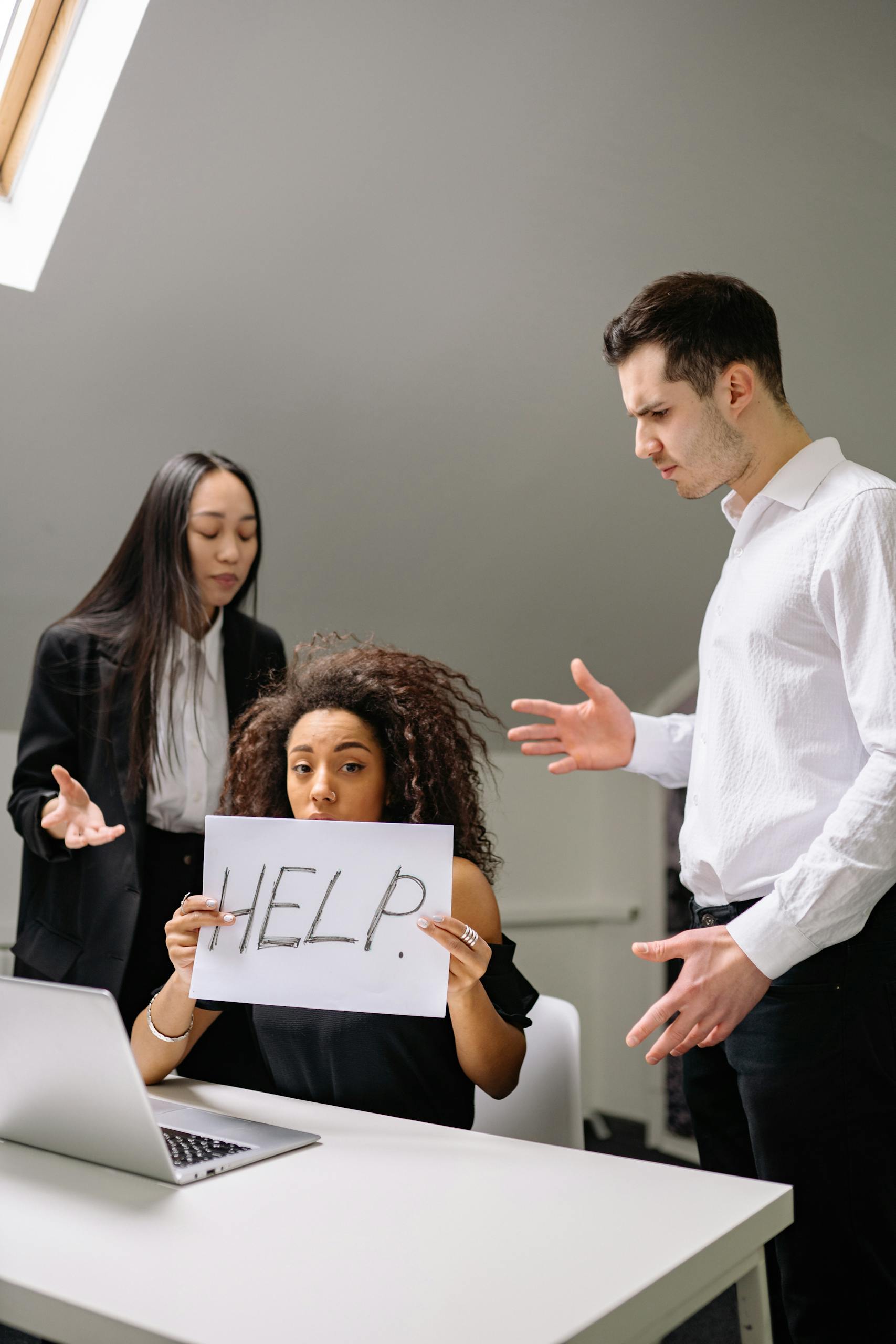 Woman holding HELP sign surrounded by colleagues in a tense office situation.